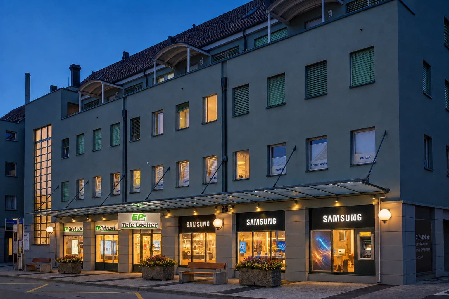 Geschäftshaus am Lindenplatz in der Dämmerung mit beleuchteten Tele-Locher- und Samsung-Schaufenstern